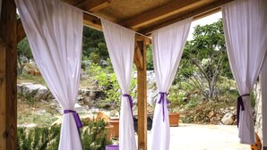 Un'area esterna riparata con tende bianche ornate da nastri viola, travi in legno e vista su un paesaggio roccioso e verdeggiante.