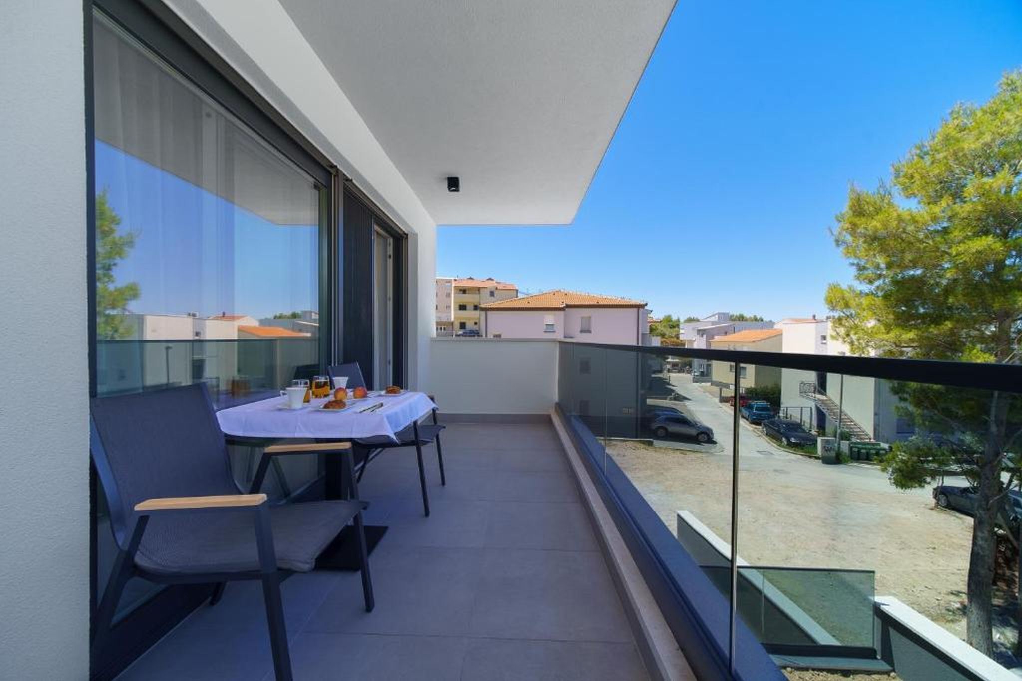 A balcony with a table and chairs, overlooking a street with parked cars and buildings.