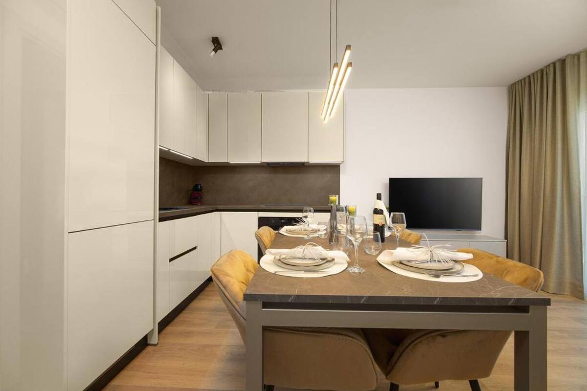 A modern dining area with a rectangular table set for four, a flat-screen TV mounted on the wall, and a kitchen area with white cabinets and a dark countertop.