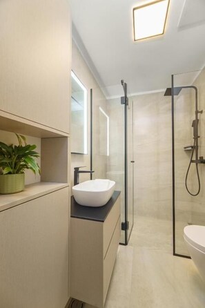 A modern bathroom with a large mirror, a white sink, a glass-enclosed shower, and a potted plant. - Apartments Bila (Podstrana)