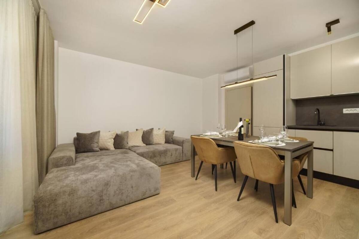 A modern living room with a grey sectional sofa, a dining table with chairs, and a kitchen area with cabinets and a countertop.