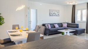 A modern living room with a grey sofa, a white coffee table, and a dining area with a white table set for four. - Apartments Nikol (Okrug)