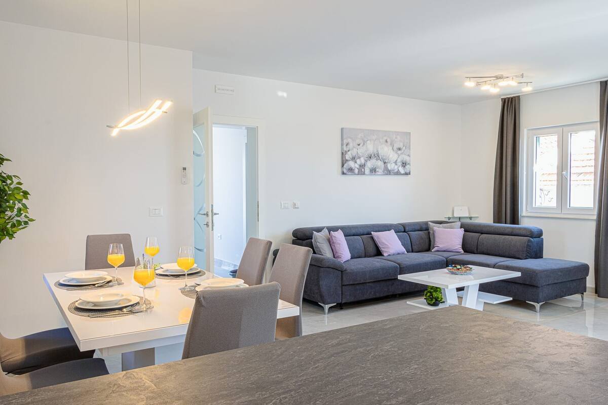 Ein modernes Wohnzimmer mit einem grauen Sofa, einem weißen Couchtisch und einem Essbereich mit einem weißen Tisch, gedeckt für vier Personen.