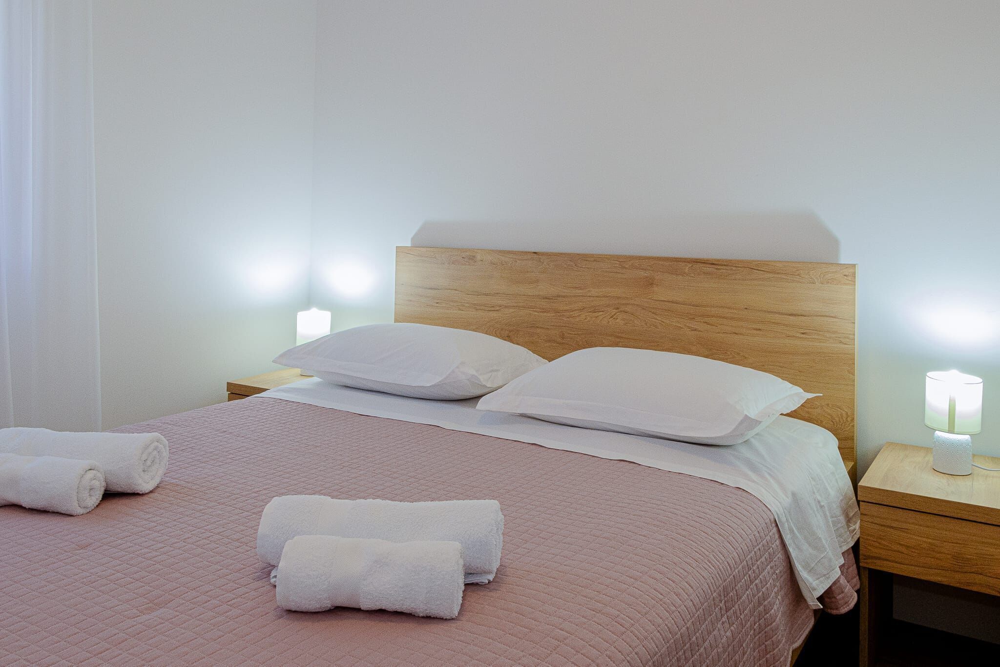 A neatly made bed with white linens and rolled towels, a wooden headboard, and bedside lamps.
