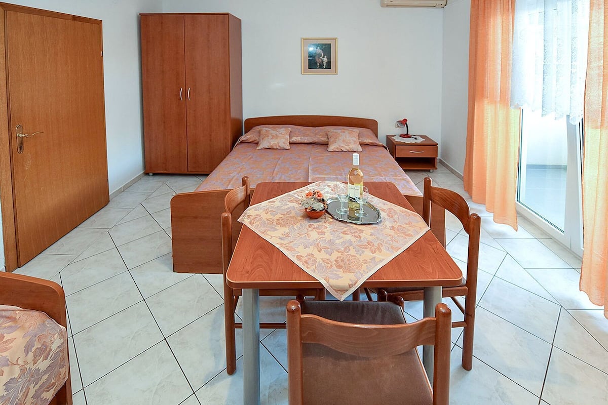 A room with a bed, a wooden wardrobe, a dining table with chairs, and a small table with a bottle and glasses.