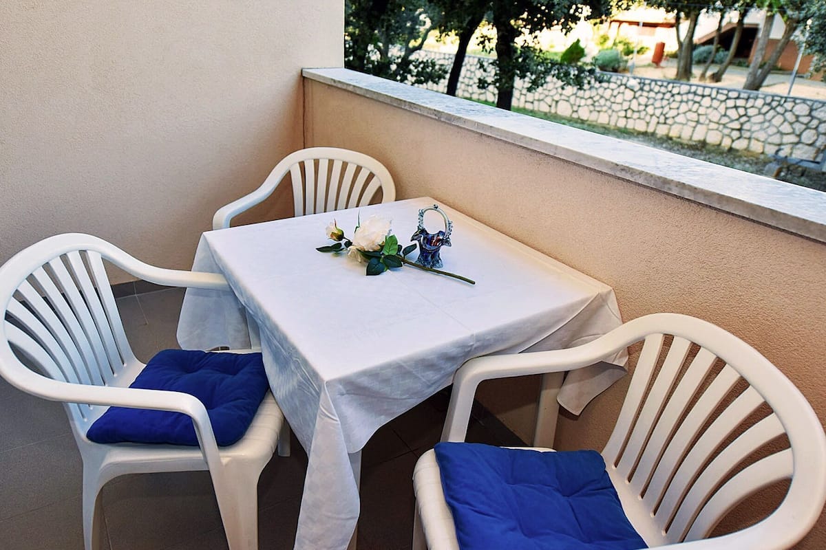 A small balcony with a table set for two, white plastic chairs with blue cushions, and a decorative vase with flowers.