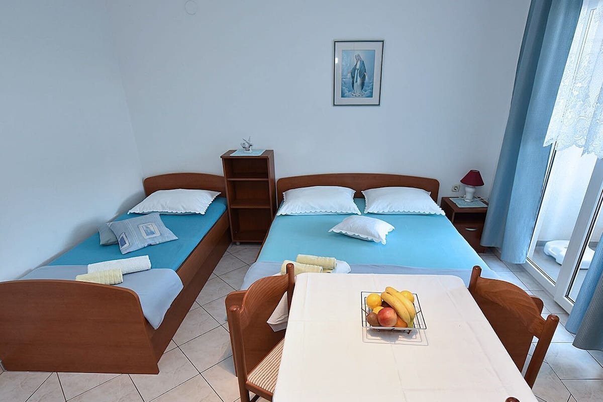 A hotel room with two single beds, a small table with a fruit bowl, a nightstand with a lamp, and a window with curtains.