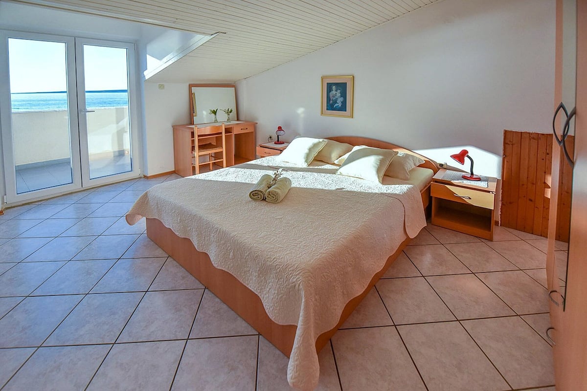 A bedroom with a large bed, a nightstand, a wooden dresser, and a window with a view of the sea.