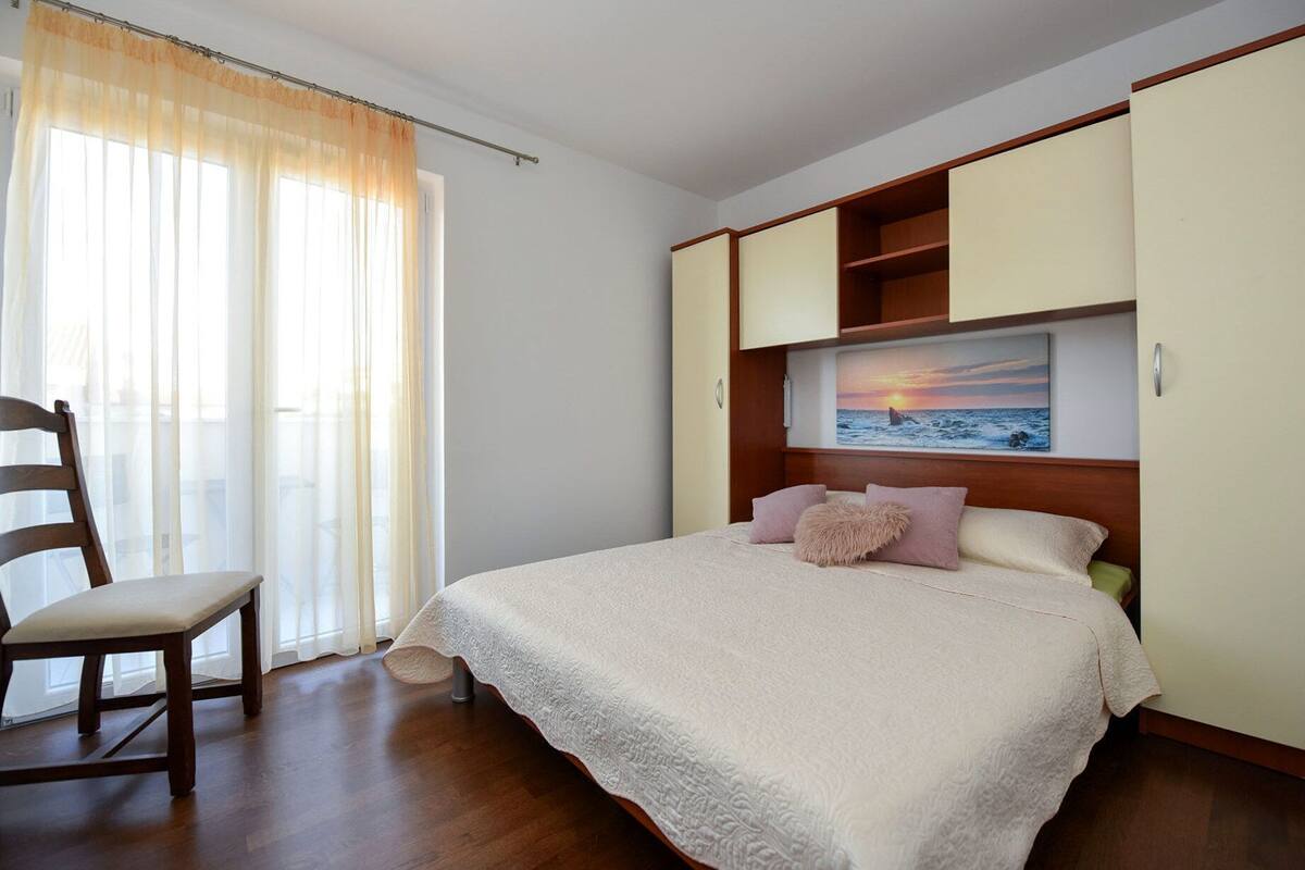 A bedroom with a bed, a chair, a painting of a sunset, and a window with sheer curtains.