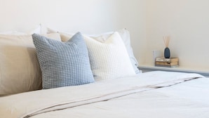 Una cama bien hecha con un edredón blanco y dos almohadas, una mesita de noche con un libro y un florero, y un aire acondicionado instalado en la pared.