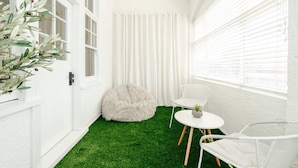 Una habitación estrecha con paredes blancas, suelo de césped artificial verde, un pouf blanco mullido, una silla blanca y una pequeña mesa con una planta.