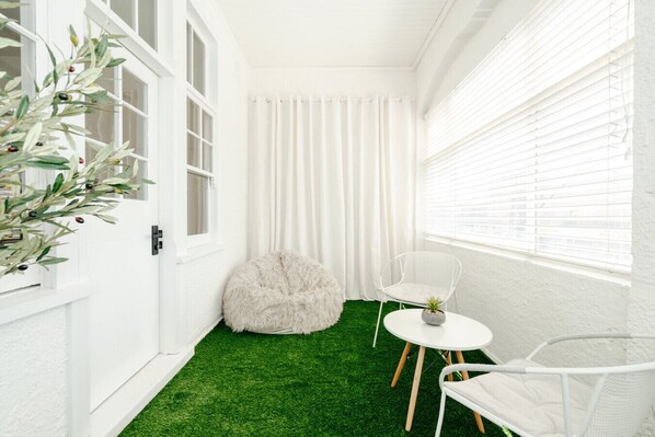 A narrow room with white walls, a green artificial turf floor, a fluffy white pouf, a white chair, and a small table with a plant.