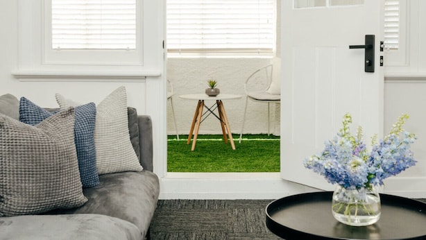 Una sala de estar con un sofá gris, una mesa de centro redonda con un florero y una puerta que da a un balcón con una pequeña mesa y una silla.