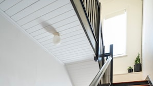 Una escalera con escalones de madera y barandillas negras, que conduce a un rellano con techo blanco y una ventana.