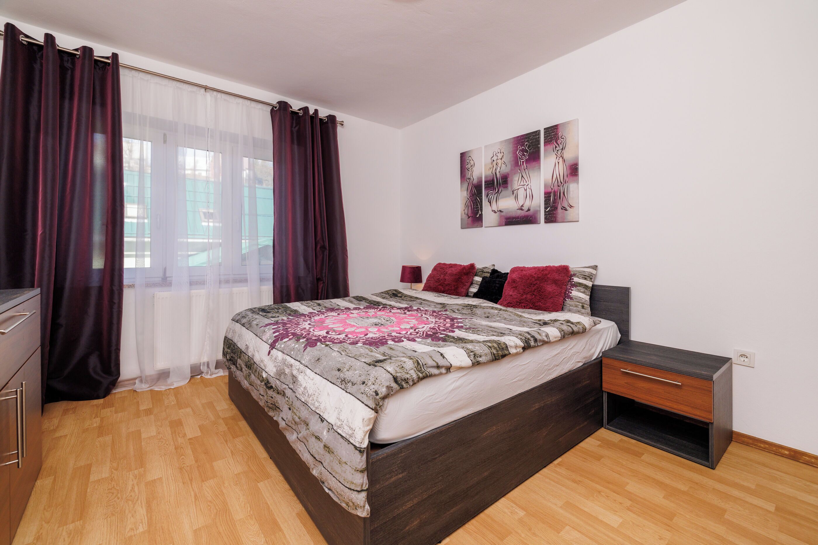 A bedroom with a wooden floor, a bed with a patterned bedspread, a nightstand, a dresser, and a window with curtains.