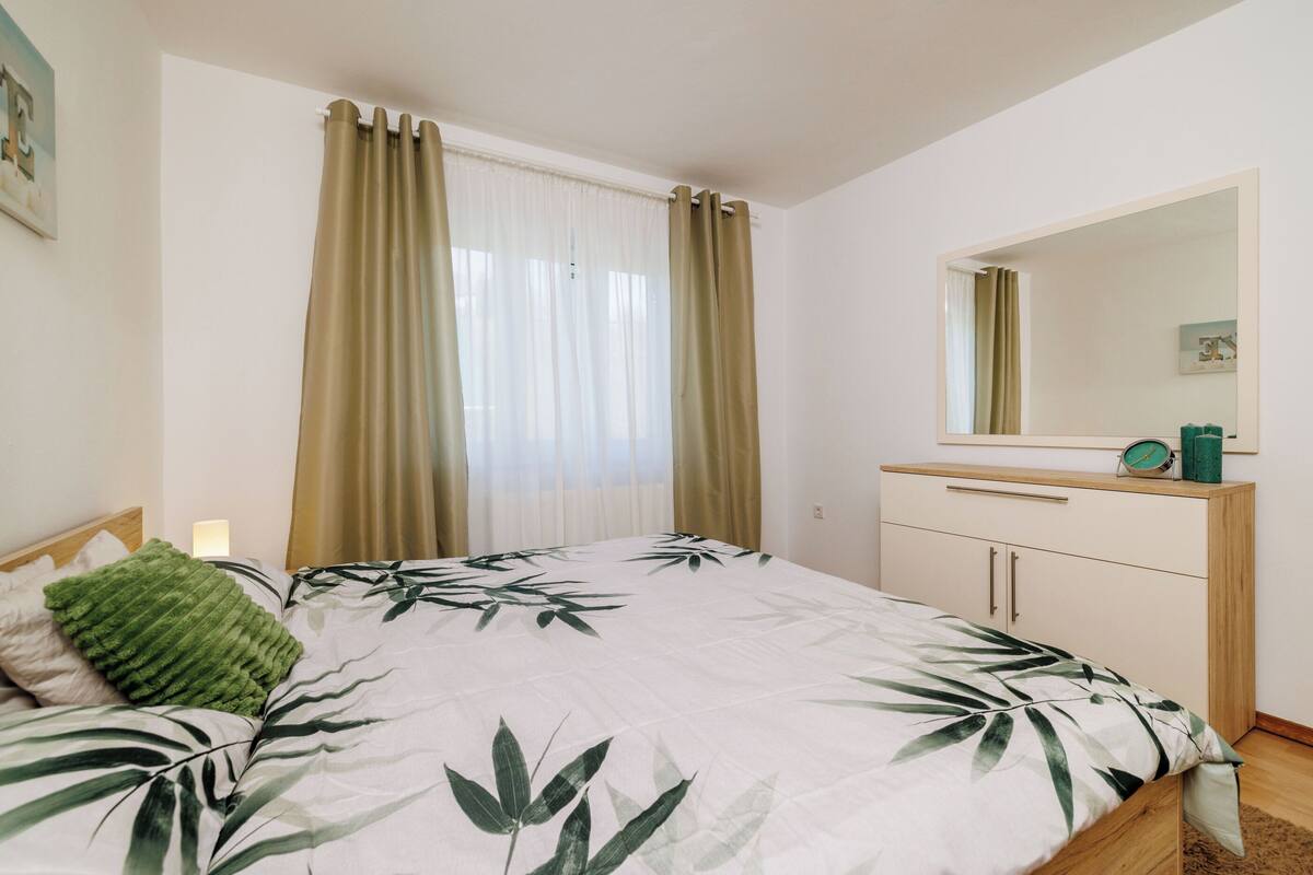 Un dormitorio con cama, mesita de noche, cómoda, espejo y ventana con cortinas.
