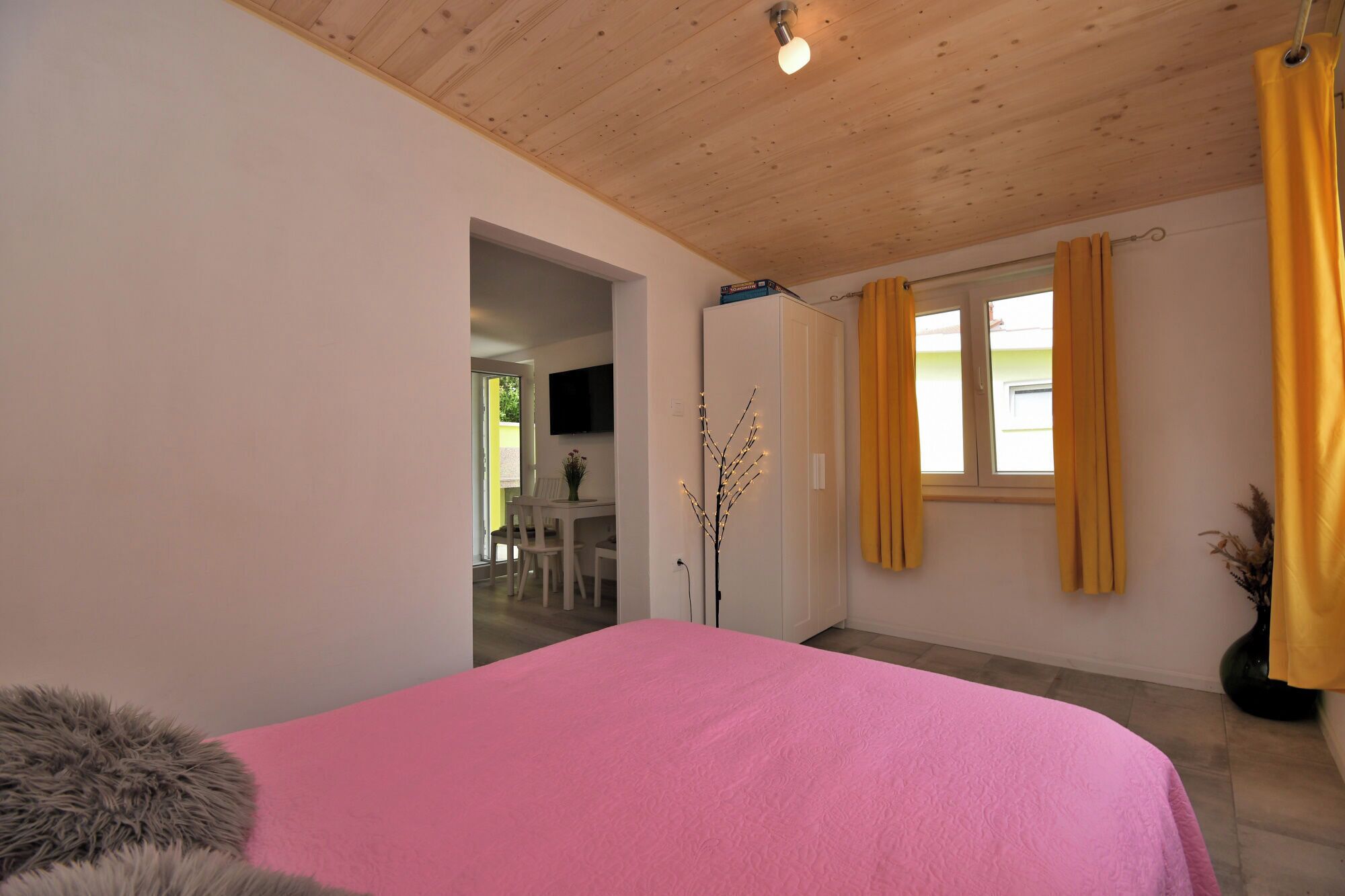 Une chambre avec un lit rose, un plafond en bois, un téléviseur fixé au mur et une fenêtre avec des rideaux jaunes.