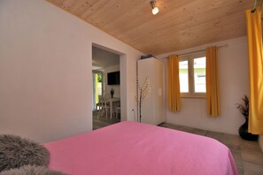 A bedroom with a pink bed, wooden ceiling, a TV mounted on the wall, and a window with yellow curtains. - Apartment Cozy Garden (Fazana)