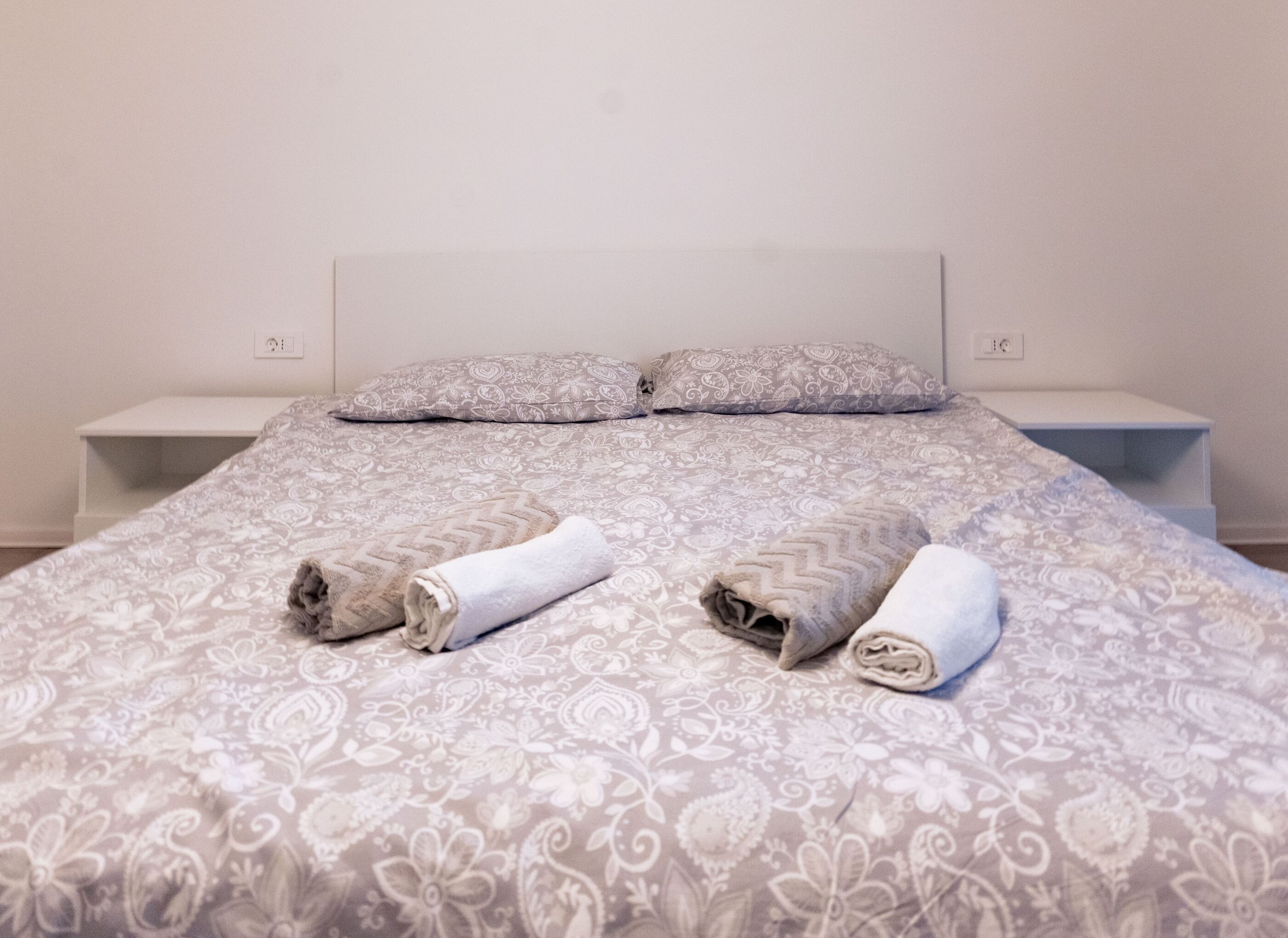 A neatly made bed with patterned bedding, two pillows, and rolled towels on a white nightstand.