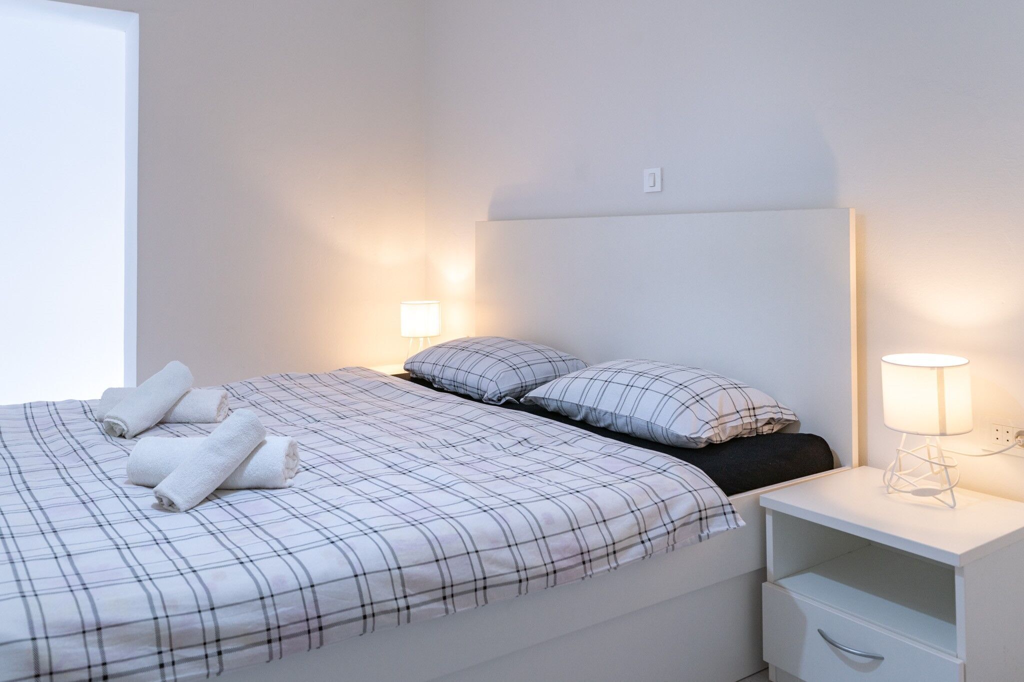 A neatly made bed with plaid bedding, a bedside table with a lamp, and a wall socket.