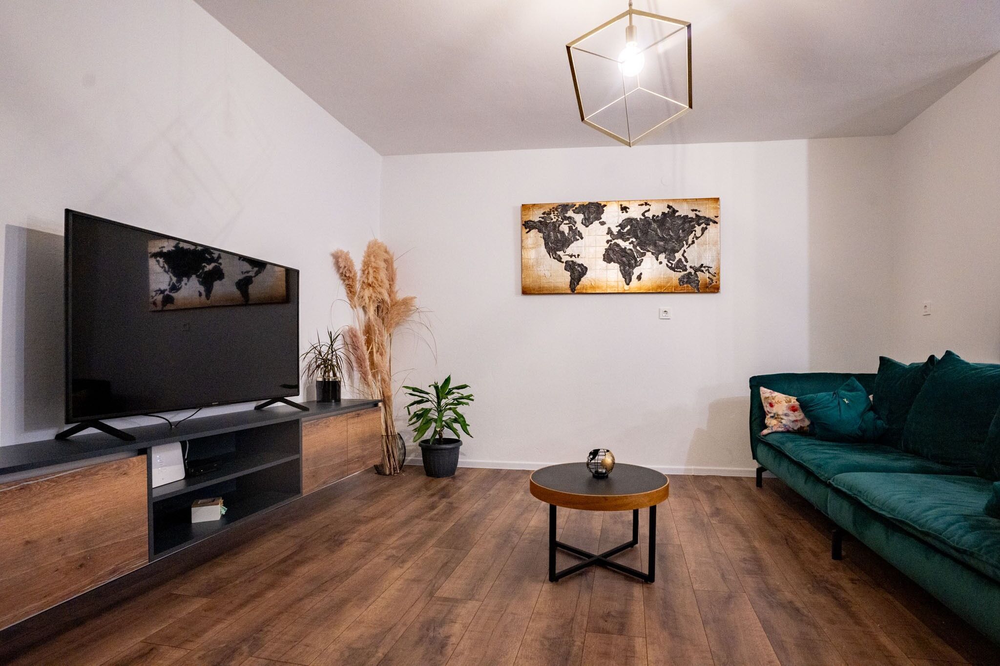 A modern living room with a flat-screen TV, a round coffee table, a green sofa, and two world map paintings on the wall.
