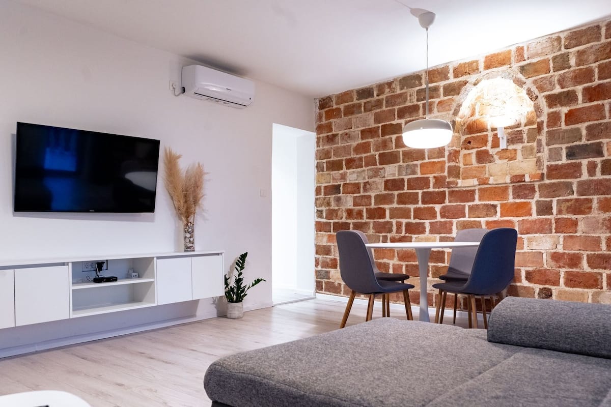 A modern living room with a flat-screen TV, a white entertainment unit, a dining area with a brick wall, and a grey sofa.