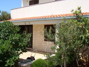 A modern building with a tiled roof, a brick wall, and a small garden area with a lawnmower and greenery. - Apartments Nadica (Rogoznica)