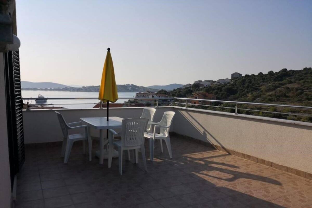 Een terras met een tafel en stoelen, een gele parasol en uitzicht op zee en heuvels.