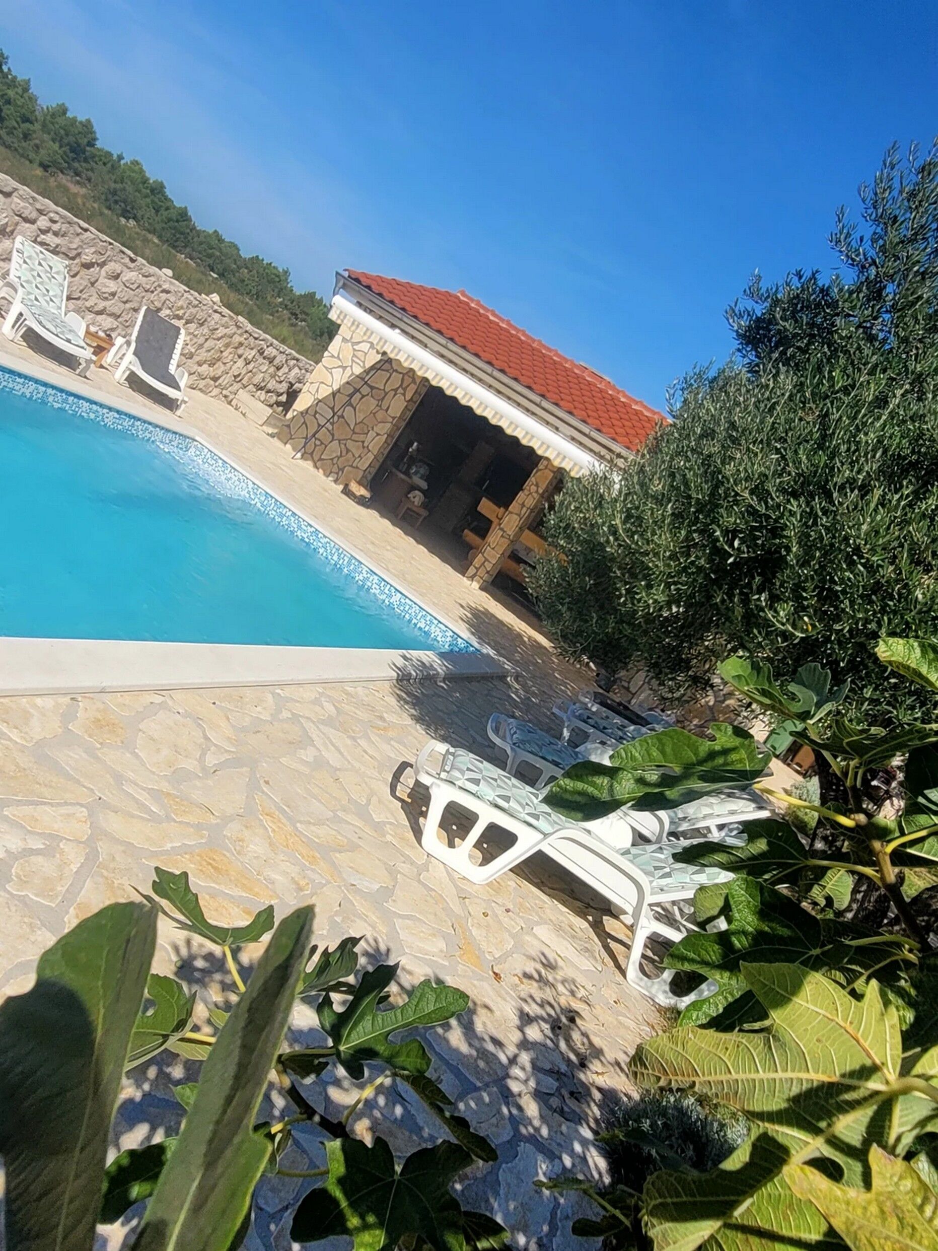 Una piscina con piastrelle in pietra, poltrone da relax bianche e un edificio in pietra con tetto rosso.