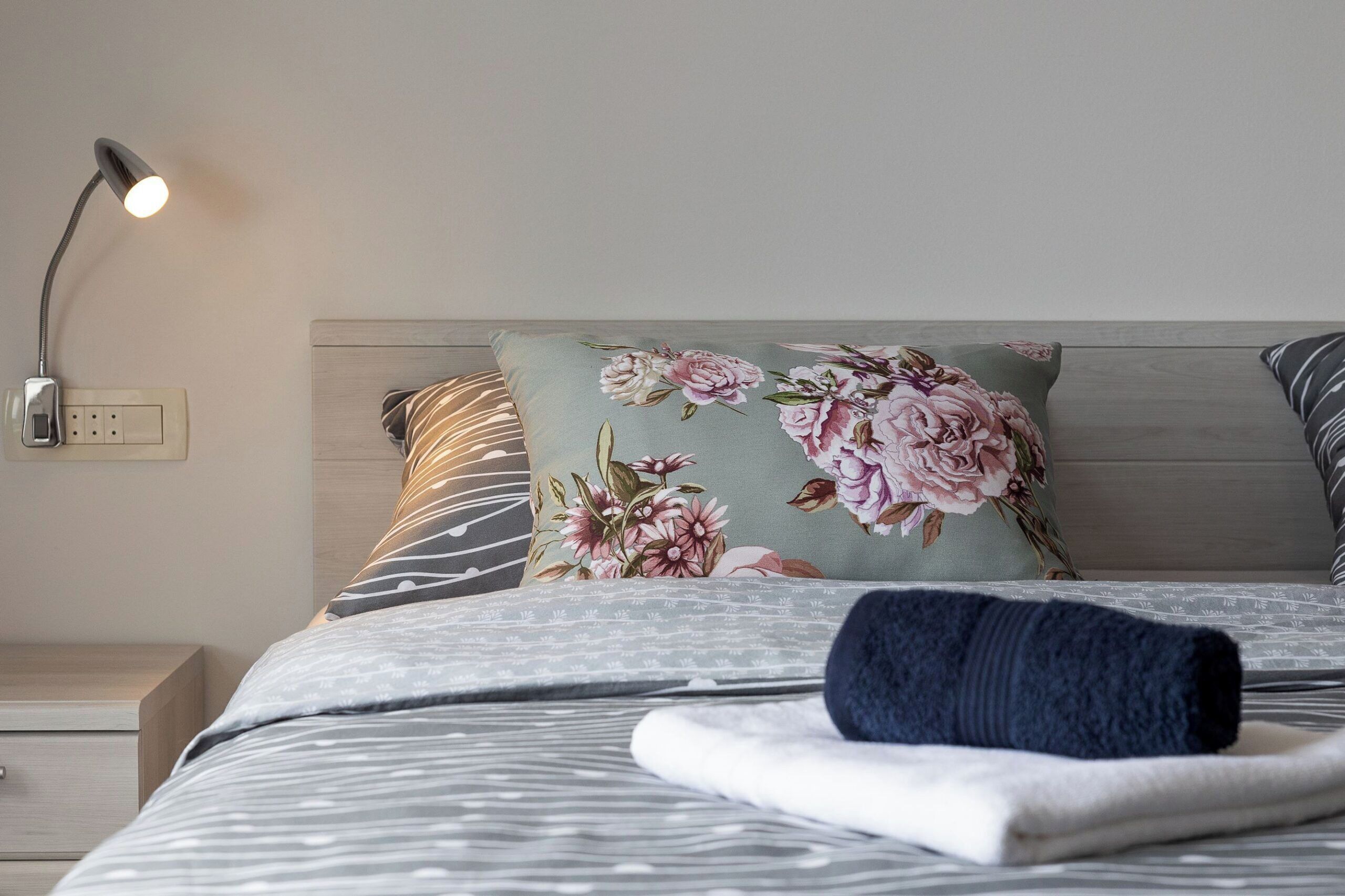 Un lit bien fait, agrémenté d’oreillers à motifs floraux, une table de chevet avec une lampe et une prise électrique au mur.