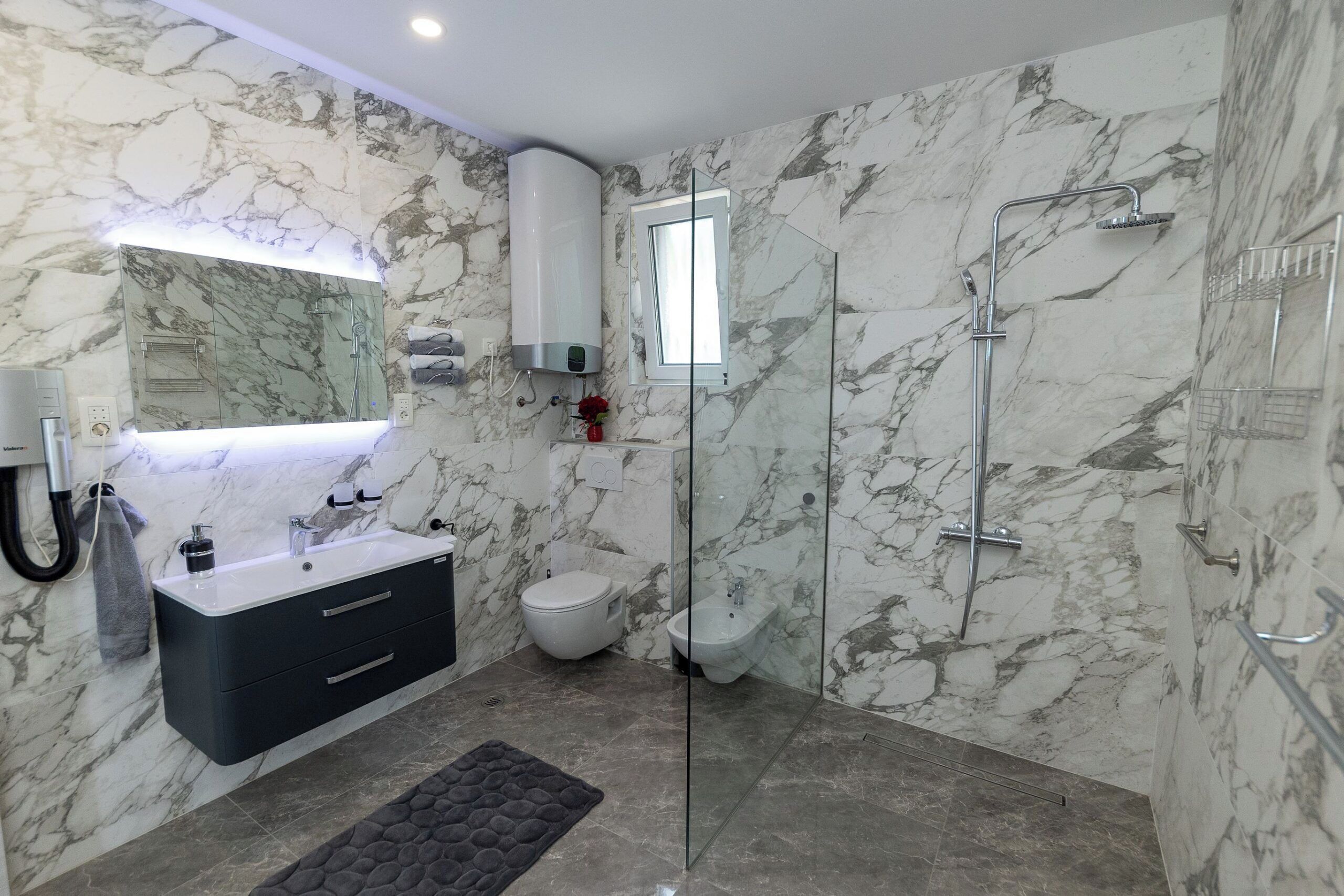 A modern bathroom with a marble wall, a glass-enclosed shower, a black vanity, and a toilet.