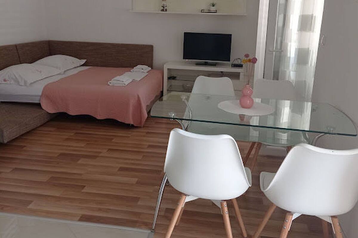 A modern bedroom with a bed, a TV, a glass dining table with chairs, and a small white cabinet.