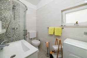 Un bagno moderno con doccia vetrata, lavandino bianco, water e finestra sopra il lavandino.