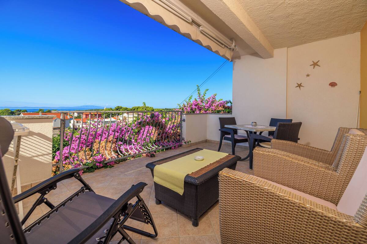 Un balcon agrémenté de meubles en osier, d’une table et de chaises, donnant sur un jardin où s’épanouissent des fleurs roses.