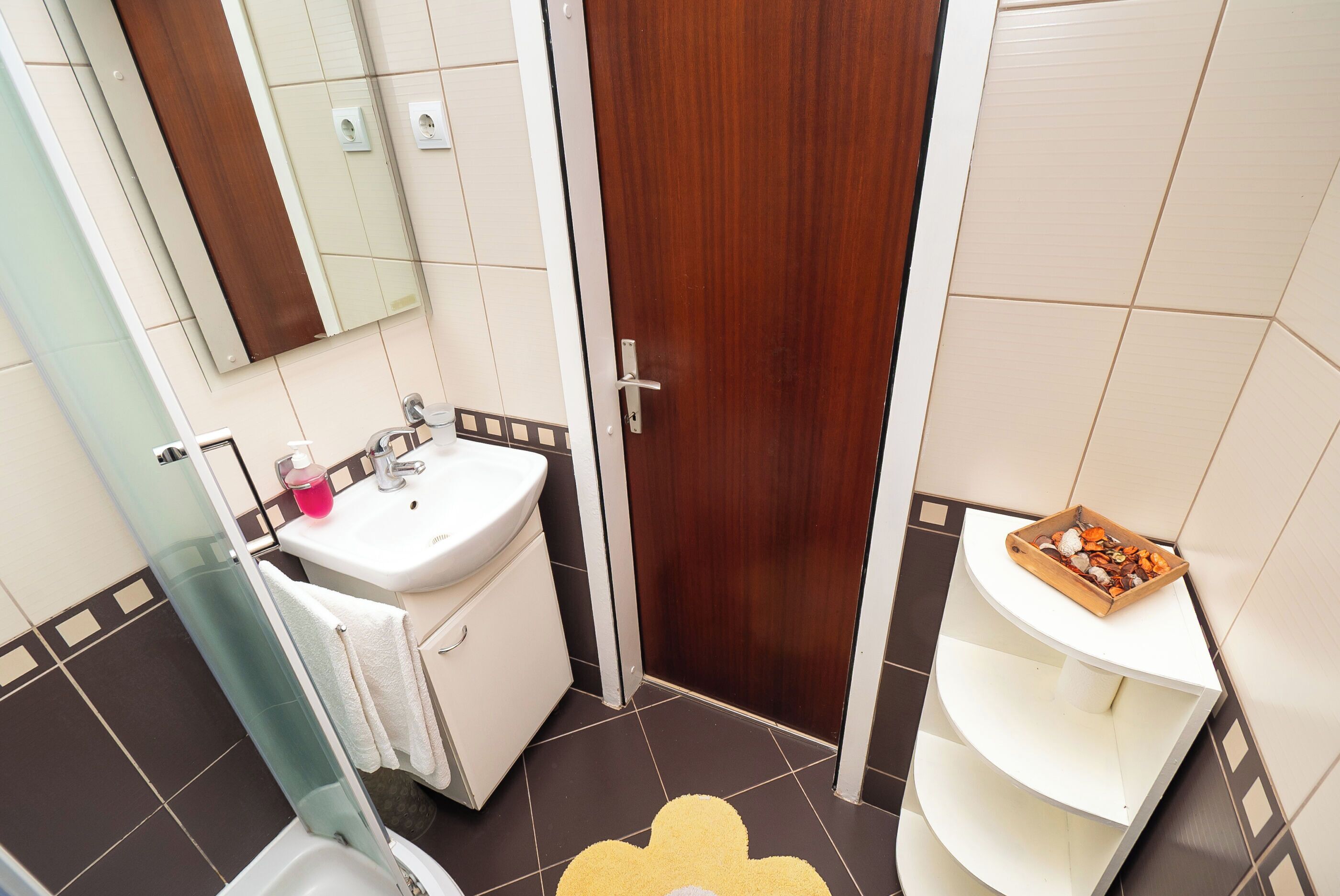 A bathroom with a white sink, a mirror, a wooden door, and a white shelf with toiletries.