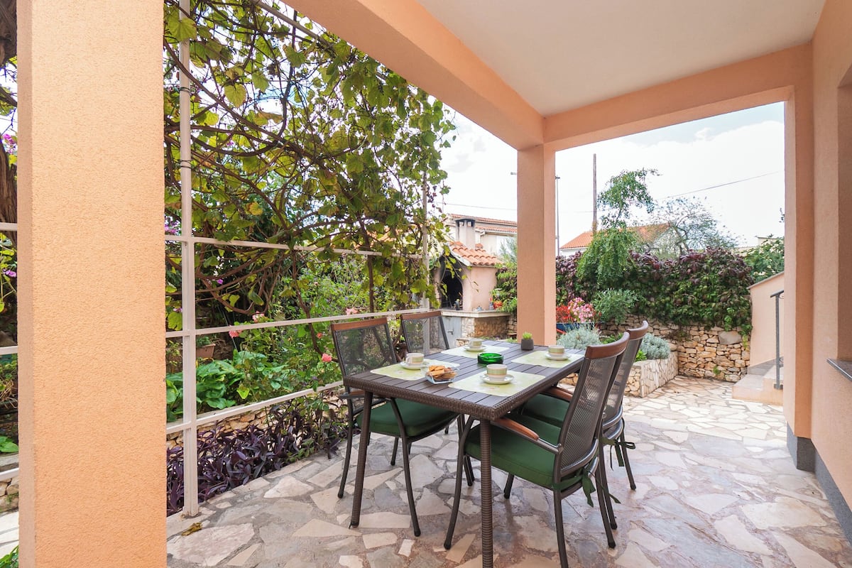 An outdoor patio with a table set for four, surrounded by green cushioned chairs, a stone floor, and a view of a garden with various plants and a trellis.