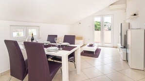 Ein moderner Essbereich mit einem weißen Tisch und violetten Stühlen, ein kleiner weißer Tisch mit einer Vase und eine Glastür zum Balkon.