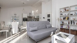 Ein modernes Wohnzimmer mit einem grauen Sofa, einem Essbereich mit einem Glastisch und einem Bücherregal, das mit Büchern und Dekorationsgegenständen gefüllt ist.