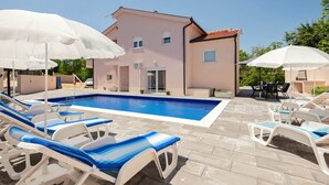 Ein Schwimmbeckenbereich mit Liegestühlen und Sonnenschirmen, ein rosafarbenes Gebäude und eine Terrasse mit Gartenmöbeln.