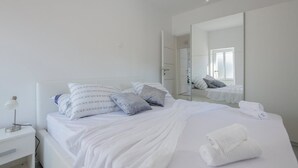 Ein ordentlich bezogenes Bett mit weißer Bettwäsche und Kissen, eine Nachttischlampe und ein zusammengerolltes Handtuch auf dem Bett.