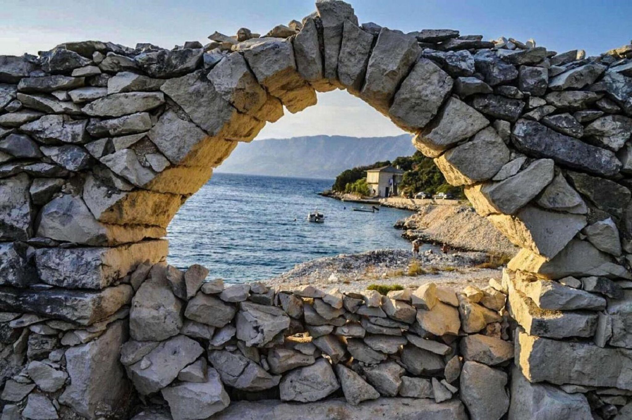 A stone archway leading to a coastal area with a view of the sea and distant mountains.