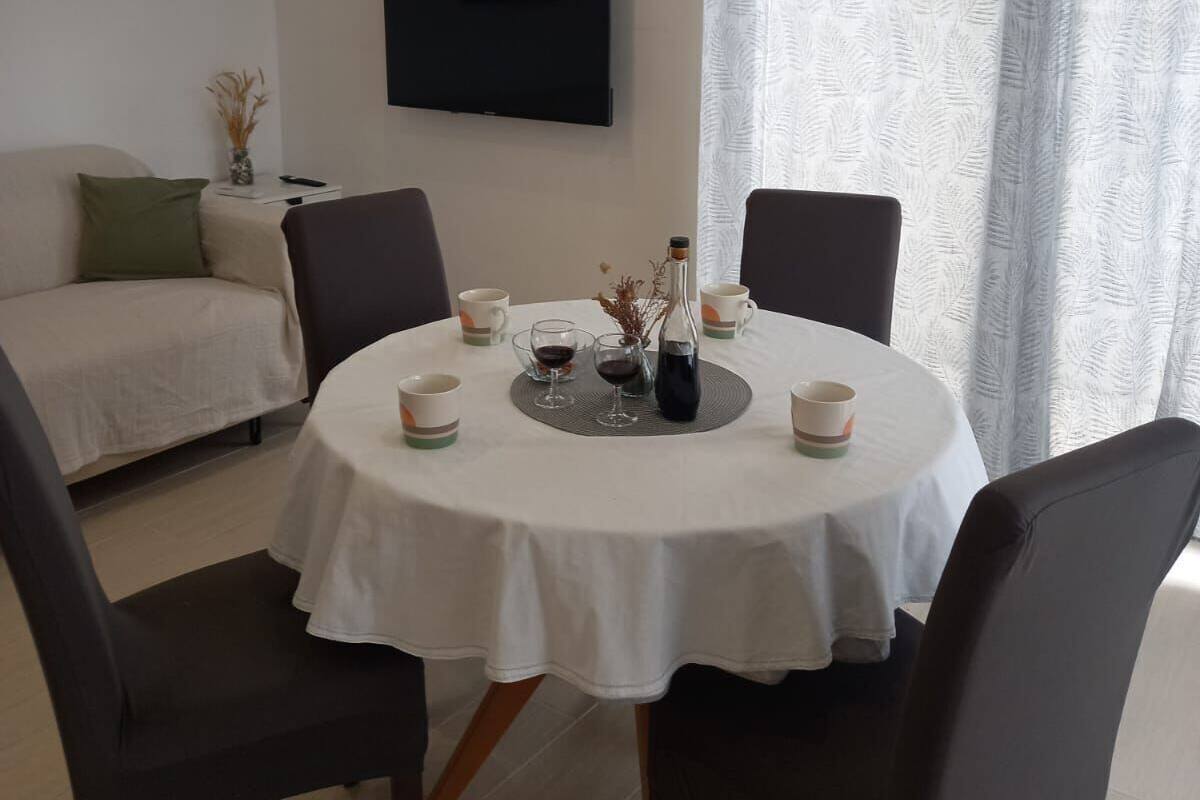 A dining area with a round table, chairs, a TV mounted on the wall, and a window with curtains.