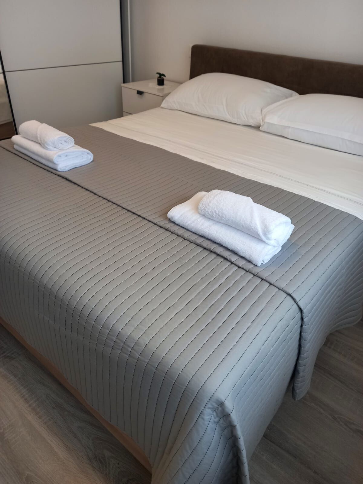 A double bed with a grey striped bedspread, two rolled white towels on the bed, a wooden headboard, and a nightstand with a small plant.
