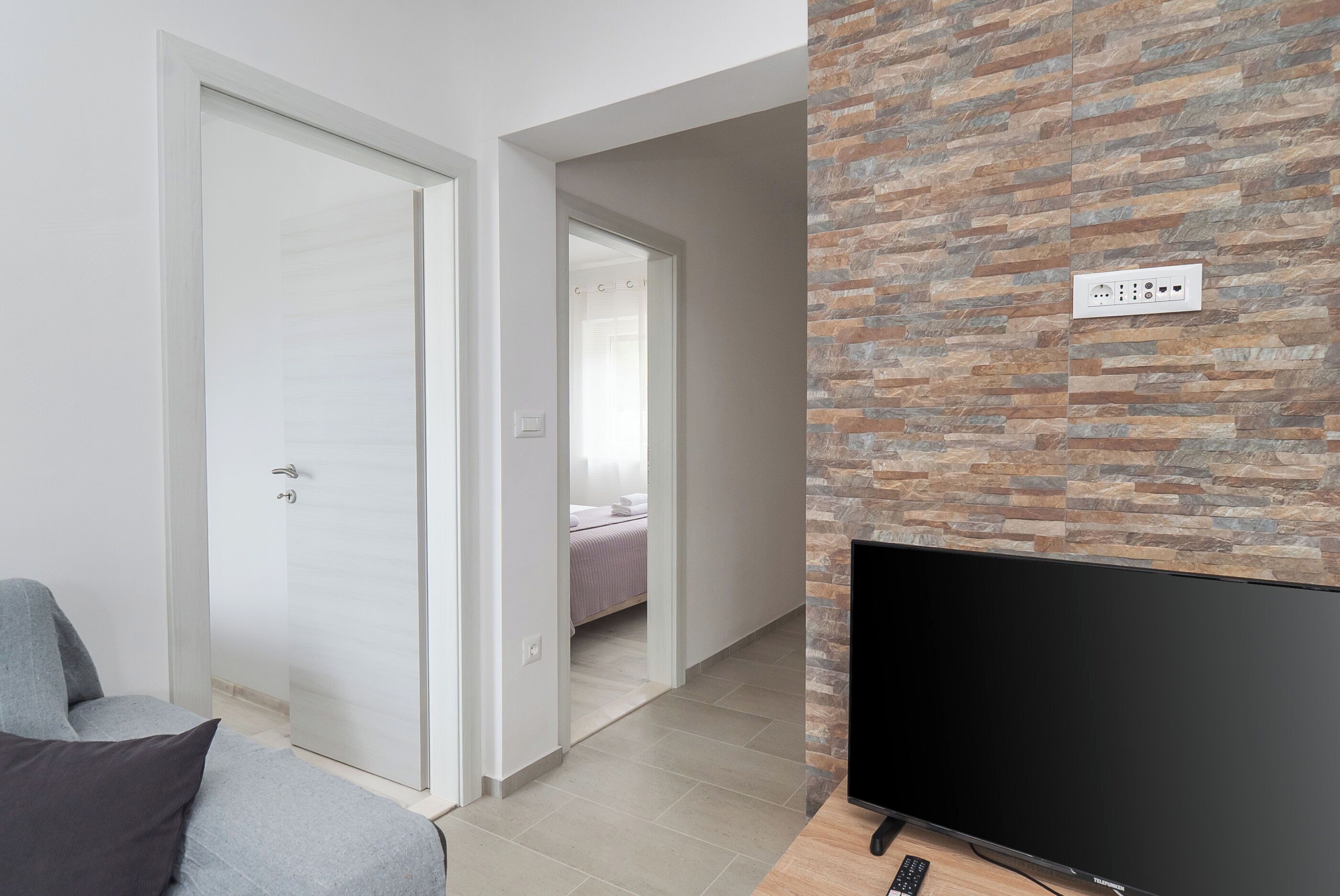 Ein modernes Wohnzimmer mit Flachbildfernseher, grauem Sofa und Wand mit Steinoptik.
