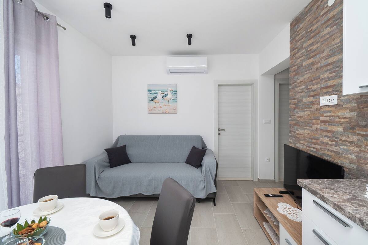 A compact living space with a grey sofa, a small dining table with cups and a bowl of fruit, a TV, and a fireplace.