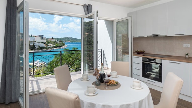 A modern kitchen with a view of the sea, a dining area with a table set for two, and a balcony with a glass door. - Apartments Kate (Rogoznica)