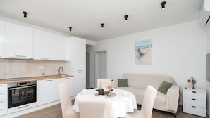 A modern kitchen with white cabinets, a built-in oven, and a dining area with a round table and chairs. - Apartments Kate (Rogoznica)