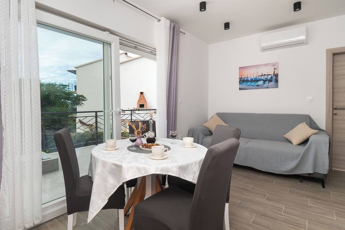 A dining area with a table set for two, a grey sofa, and a view of a building with a chimney.
