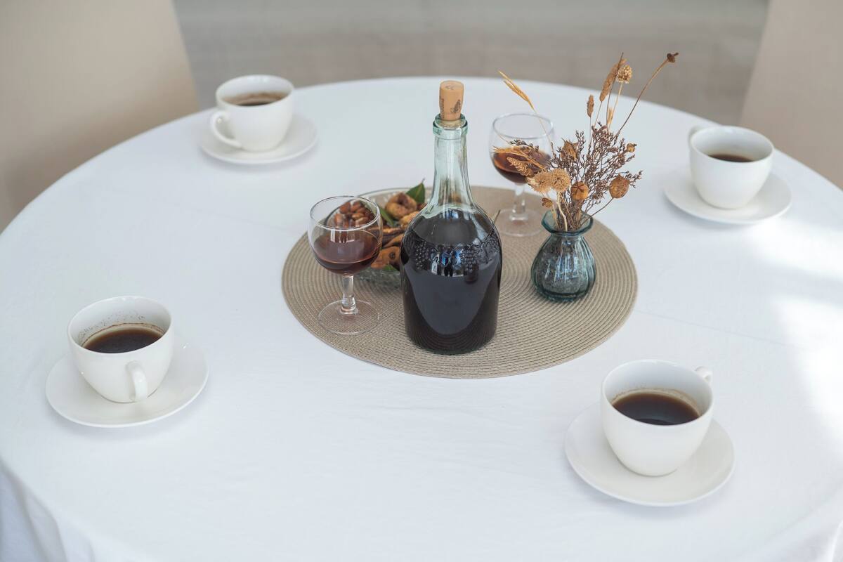 A round table with a bottle of wine, a vase with dried flowers, and four cups of coffee.