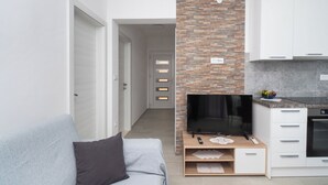 A modern living room with a grey sofa, a flat-screen TV on a wooden stand, and a kitchen area with white cabinets and a stone backsplash. - Apartments Kate (Rogoznica)